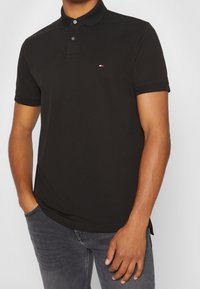 Polo noir avec col, deux boutons et un petit logo sur la poitrine ; fabriqué en tissu texturé, associé à un jean gris.