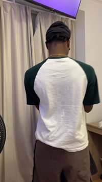 T-shirt with green raglan sleeves and a white body, paired with brown textured pants. Background includes curtains and furniture.