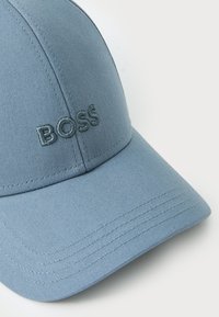 Lys blå baseballcaps laget av teksturert stoff med et brodert logo "BOSS" på forsiden. Har en buet skygge og sekspaneldesign.