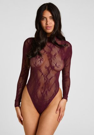 Femme aux longs cheveux foncés portant un body en dentelle bordeaux transparent à manches longues sur un fond gris uni.