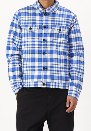 Mężczyzna ubrany w niebiesko-białą kratowaną kurtkę zapinaną na guziki z dwoma kieszeniami na piersi oraz czarne spodnie, ręce swobodnie opuszczone wzdłuż ciała.