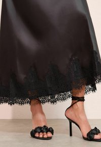 Abito di satin nero con dettagli in pizzo lungo l'orlo; sandali neri con tacco alto decorati con rose in tessuto e sottili cinturini alla caviglia.