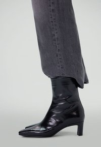 Bottes noires à chevilles avec une finition laquée brillante, bout pointu et un talon sculpté, associées à un jean gris fuselé montrant une légère effilochure à l'ourlet.