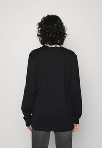Camisa preta de manga longa com decote redondo, apresentando um colarinho estampado. Textura suave e corte descontraído, combinada com calças cinzentas.