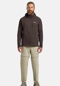 Giacca marrone con cappuccio e cerniera, tessuto texturizzato; tasca sul petto con cerniera e due tasche frontali. Pantaloni beige chiaro con cuciture.