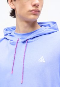 Νεαρό άτομο που φοράει λουλακί φούτερ με κουκούλα και ροζ κορδόνια, με ένα μικρό λευκό λογότυπο ACG στο στήθος, κοιτάζοντας λίγο προς τα δεξιά.