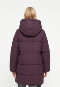 Lilla puffjakke med hette, med quilte-seksjoner og en avslappet passform. Materialet ser glatt og isolert ut for varme.