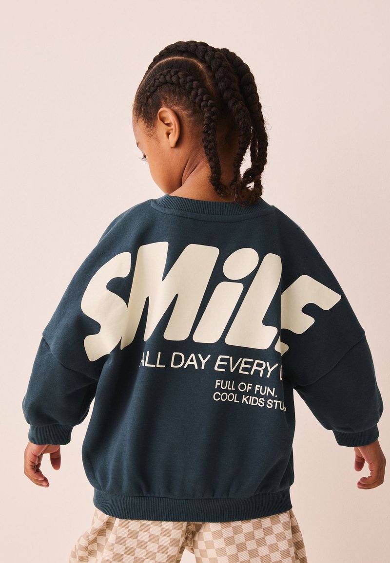 Niño con trenzas que lleva una sudadera azul oscuro con un gran texto blanco "SMILE" y pantalones a cuadros, mirando de espaldas a la cámara.