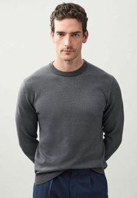 Grauer Rundhalspullover mit glatter Textur, gerippten Bündchen und Saum. Getragen von einer Person, die mit verschränkten Händen steht und mit einer marineblauen Hose kombiniert ist.