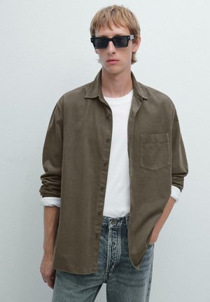 Camicia verde oliva con bottoni e taschino sul petto, abbinata a una maglietta bianca e jeans in denim chiaro, modello che indossa occhiali da sole neri.