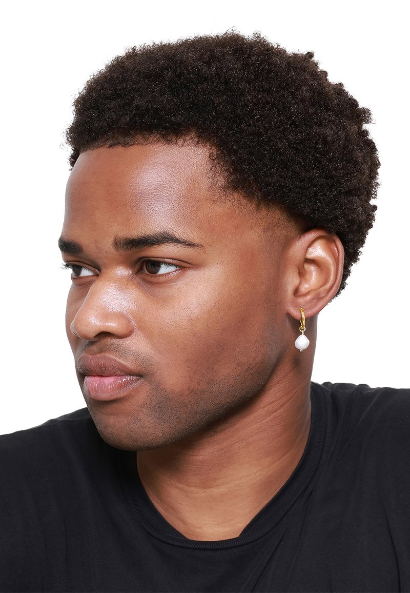 Boucle d'oreille en or avec un accent en perle blanche. Le modèle a les cheveux noirs courts et bouclés et porte une chemise noire. Vue de profil.