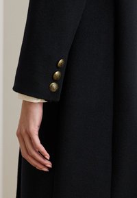 Μαύρο μάλλινο παλτό με ραφτό σχέδιο, διακοσμημένο με τέσσερα περίτεχνα χρυσά κουμπιά στο μανίκι και λεία υφή.