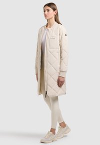 Donna in piedi che indossa un lungo cappotto trapuntato beige, leggings crema, sneakers bianche e una maglietta bianca su uno sfondo grigio uniforme.