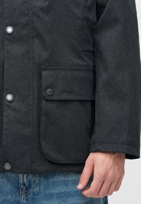 Close-up of a person wearing a dark grey Barbour jacket with metallic buttons and a large front pocket, paired with blue denim jeans.