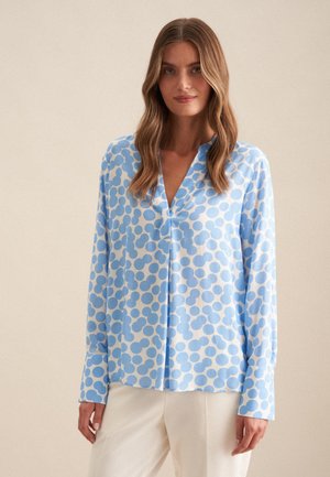 Lichtblauwe blouse met een wit cirkelpatroon, een V-hals, lange mouwen en een ontspannen pasvorm. De stof lijkt soepel en lichtgewicht te zijn.