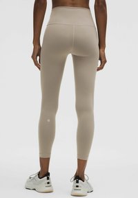 Beige high-waisted leggings met een gladde textuur en een getailleerd ontwerp, voorzien van een logo-detail nabij de enkel en gecombineerd met witte sportschoenen.