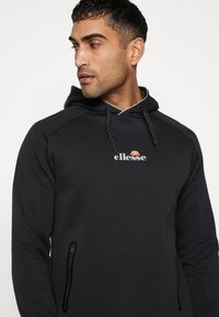Black hooded sweatshirt with a front logo. Features zippered side pockets and a textured fabric. Contrast stitching outlines the hood.