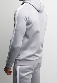 Felpa con cappuccio grigio chiaro realizzata in tessuto morbido, dotata di tasca frontale, orlo a coste e strisce laterali bianche. Texture liscia e design aderente.