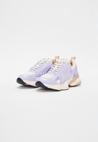Zapatillas deportivas moradas con paneles de malla, acentos beige, suelas texturizadas y detalles reflectantes. Incluyen cordones y talones reforzados.