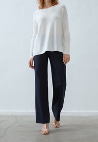 Valkoinen, teksturoitu neulepaita pitkillä hihoilla yhdistettynä tummansinisiin leveälahkeisiin housuihin. Vaalean kultaiset sandaalit näkyvät jaloissa. Yksinkertainen, klassinen muotoilu.