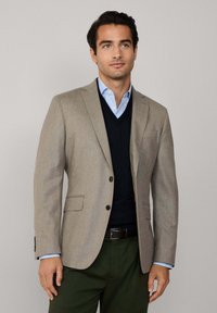 Ljusbrun blazer med två knappar, slagkrage och två framfickor. Bärs över en marinblå V-ringad tröja och en ljusblå skjorta. Gröna byxor.