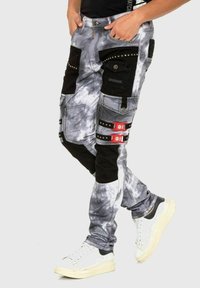 Pantalons cargo en denim, avec un motif tie-dye gris, des accents noirs, de multiples poches et des détails de boucle rouges.