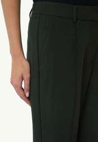 Ciemnozielone spodnie o dopasowanym kroju z gładką teksturą, z kieszeniami bocznymi i eleganckim fasonem, przedstawione z ręką blisko talii.