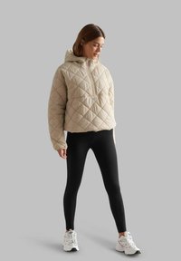 Beige quilted hooded jacket with a half-zip front, paired with ribbed black leggings and white athletic shoes.