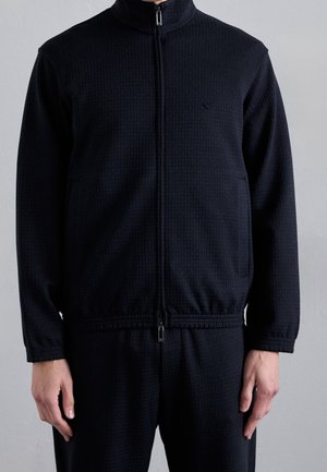 Hombre vistiendo una chaqueta texturizada azul marino oscuro con cremallera y cuello alto, junto con pantalones a juego, de pie frente a un fondo claro y liso.