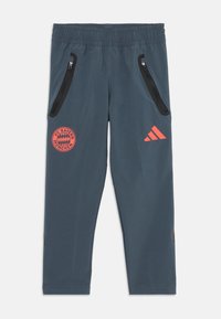 Blå treningsbukser med elastisk liv, to glidelåslommer på siden, rødt FC Bayern München-logo og oransje Adidas-detalj.