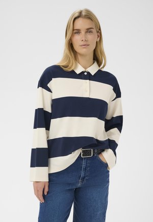 Blonde Frau trägt ein marineblau-weiß gestreiftes langärmeliges Polohemd, das in blaue Jeans mit schwarzem geflochtenem Gürtel gesteckt ist.