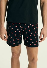 Czarne kąpielówki z wzorem różowych serc, o gładkiej fakturze i regularnym kroju. Regulowany pas bez widocznych elementów.