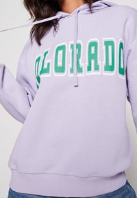 Donna che indossa un hoodie color lavanda con testo verde e bianco "COLORADO", abbinato a jeans blu, inquadrata dalla vita al petto.