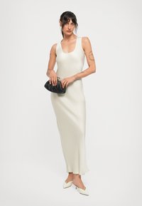Kvinna i en ärmlös vit klänning som når anklarna, hållande en svart kuvertväska, klädd i vita kitten heels, stående mot en enkel bakgrund.