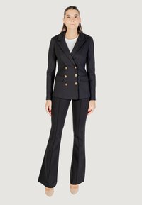 Donna in piedi che indossa un blazer doppiopetto nero con bottoni dorati, pantaloni neri a zampa d'elefante, top bianco e tacchi beige, su sfondo bianco.