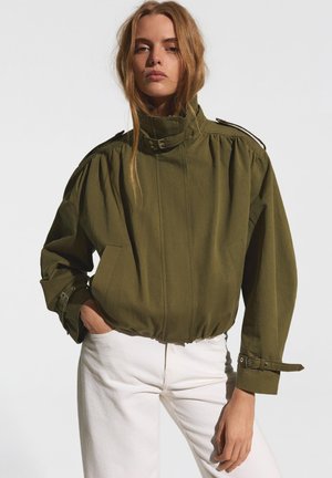 Frau trägt eine olivgrüne Jacke mit hohem Kragen und Schnallendetails sowie weiße Hosen und steht vor einem schlichten hellen Hintergrund.