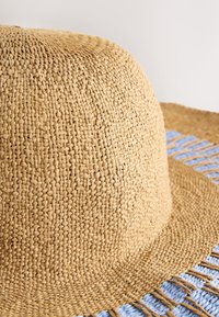 Cappello di paglia a tesa larga con una superficie marrone chiaro texturizzata e dettagli intrecciati azzurri lungo il bordo della tesa.