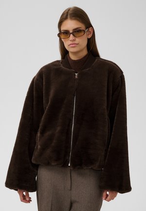 Vrouw met steil bruin haar draagt een bruine nep-bont jas met rits, een bruine trui, bruin getinte zonnebril en grijze broek, kijkt naar voren.