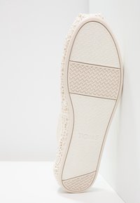 Chaussure à lacets avec une tige en crochet crème, semelle en caoutchouc texturée présentant un motif en chevrons et un léger soutien de la voûte plantaire. Logo de la marque visible.