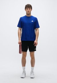 Modra športna majica s kratkimi rokavi, v kombinaciji s črnimi kratkimi hlačami. Model drži bordo elastični trak za vadbo. Bele superge s sivimi poudarki.