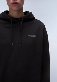 Zwarte hoodie gemaakt van zachte stof, met een klein rechthoekig logo op de borst. De koorden zijn zichtbaar op de capuchon.