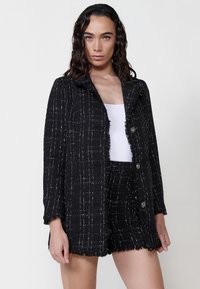 Jachetă neagră țesută cu un finisaj texturat, având o margine cu franjuri, butoane de ton argintiu și un model în grilă. Purtață peste un top alb.