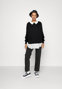 Pull noir surdimensionné avec un col blanc superposé sur une chemise blanche, associé à un jean noir et des baskets noires. Un simple chapeau noir porté.