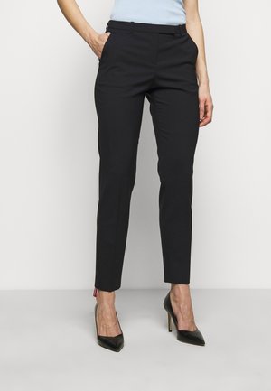 Person, die schlanke schwarze Hose und schwarze High Heels trägt, steht mit den Händen in den Taschen vor einem schlichten weißen Hintergrund.