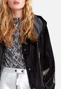 Černá lesklá bunda s kapucí, nošena přes šedý top s hadím potiskem. Stříbrné metalické kalhoty se zipovým zapínáním a cvoky.