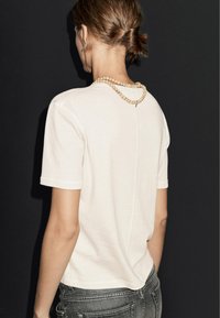 Krabelig, kortærmet t-shirt i cremefarve med en central syning, parret med en halskæde af træperler og grå jeans, set fra bagsiden.