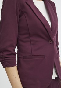 Blazer bordeaux sur mesure à un seul bouton, épaule structurée et manches retroussées. Comprend deux poches latérales et une texture lisse.