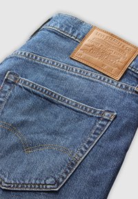 Jeans de ganga azuis com uma etiqueta de couro com texto em relevo. Detalhes de costura visíveis e bolsos traseiros arredondados. Design clássico.