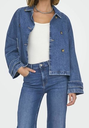 Femme portant une veste en jean bleue, un jean bleu taille haute, un top blanc et un collier chaîne, posant avec une main dans la poche.