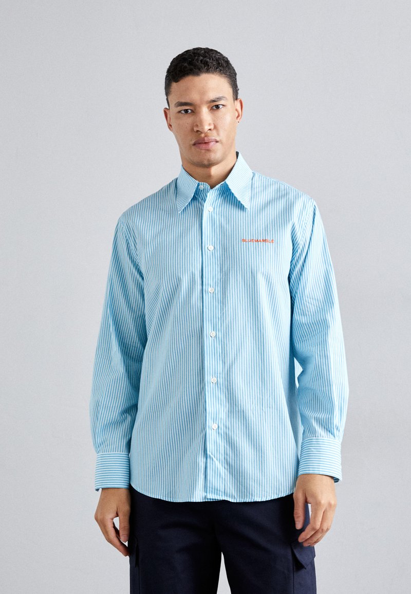 Camisa a rayas de color azul claro con un diseño de botones, que presenta un cuello puntiagudo y un pequeño logo naranja bordado en el pecho.